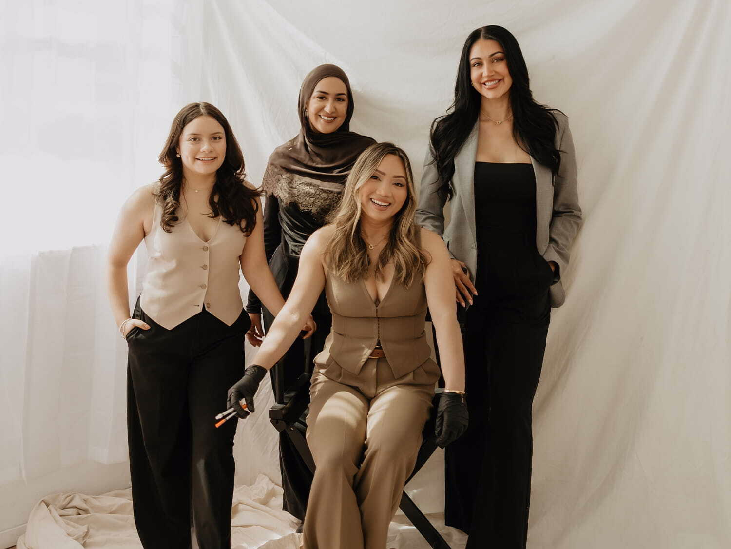 Four diverse women posing indoors, three standing and one seated, all smiling and dressed in business attire.