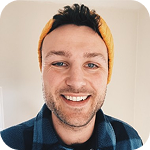 Smiling man wearing a yellow headband and a blue checkered shirt against a light wall.