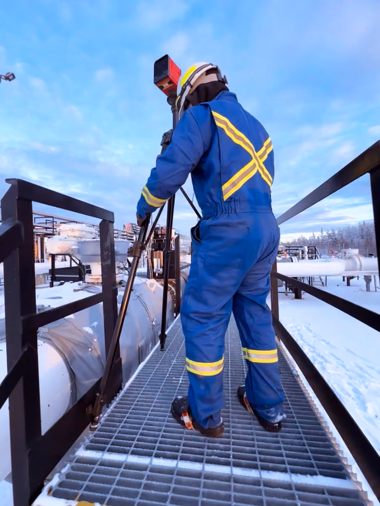 Worker in blue protective coveralls with yellow reflective stripes using surveying equipment on an industrial platform in a snowy outdoor setting during daytime.