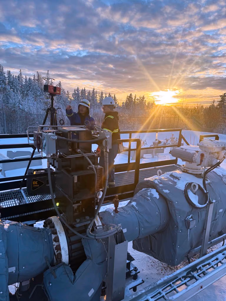 Two workers wearing helmets and safety clothing operating industrial equipment on a snowy platform during sunset with a forest background.