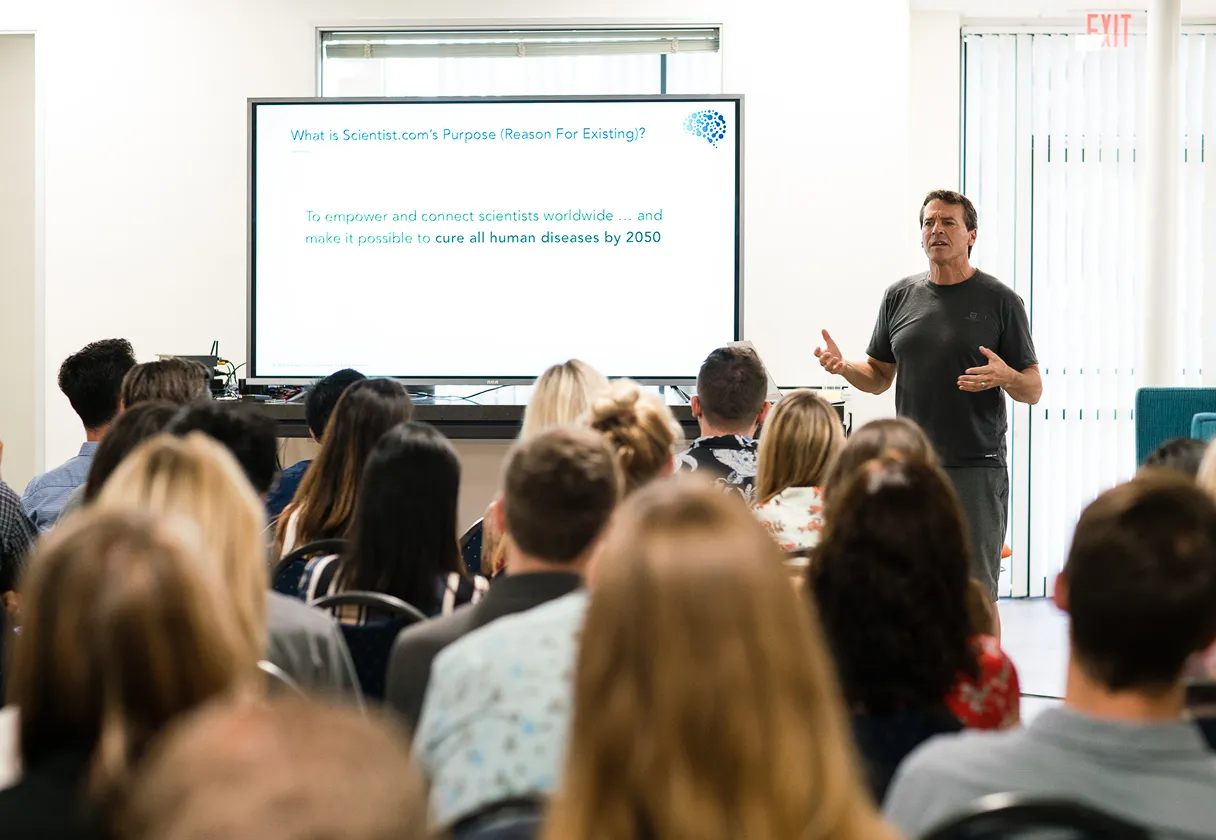 Man giving a presentation to an audience with a screen about curing all human diseases by 2050.
