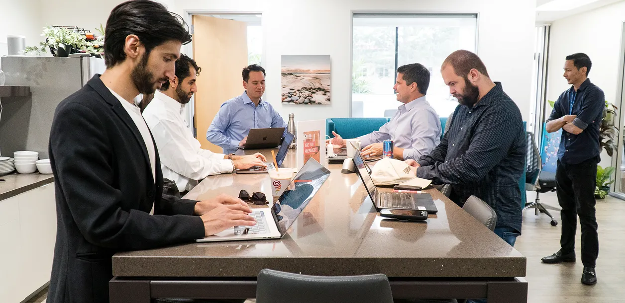 Six men working and discussing around a kitchen island with laptops in a modern office space.