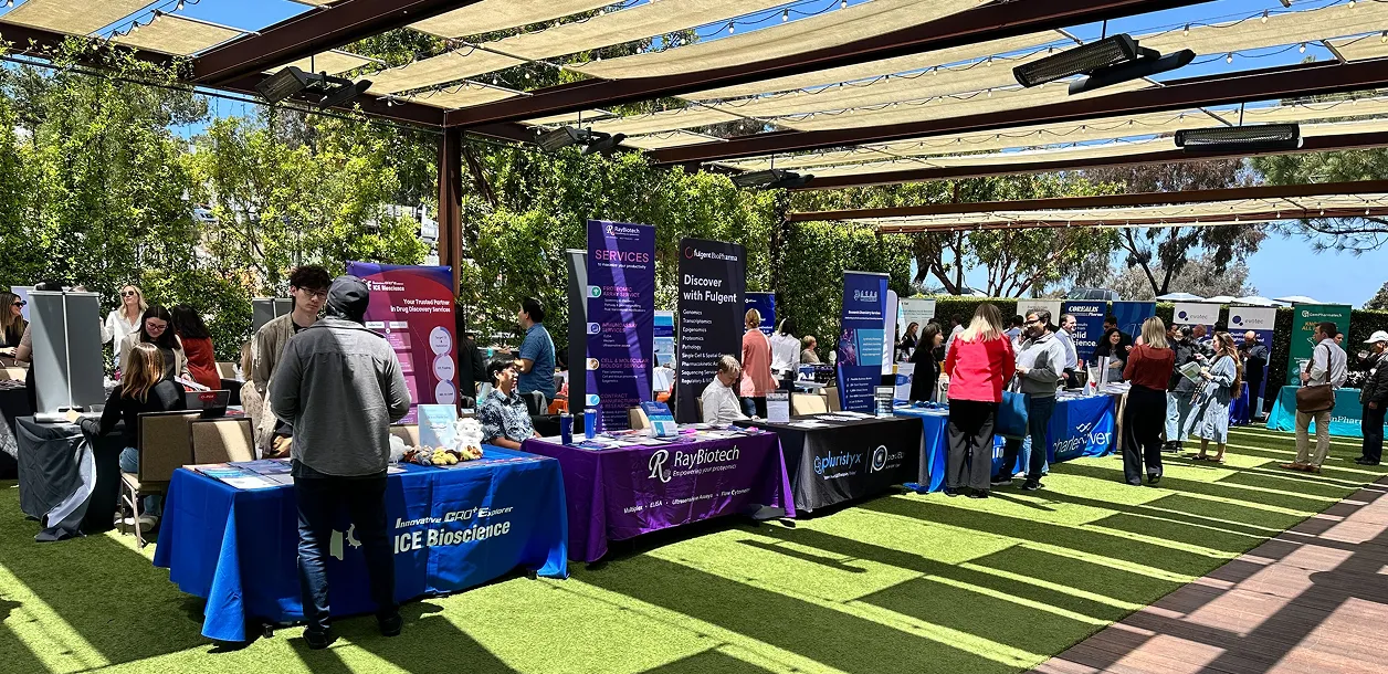 Outdoor biotech fair with multiple company booths, people engaging and discussing under shade structures.