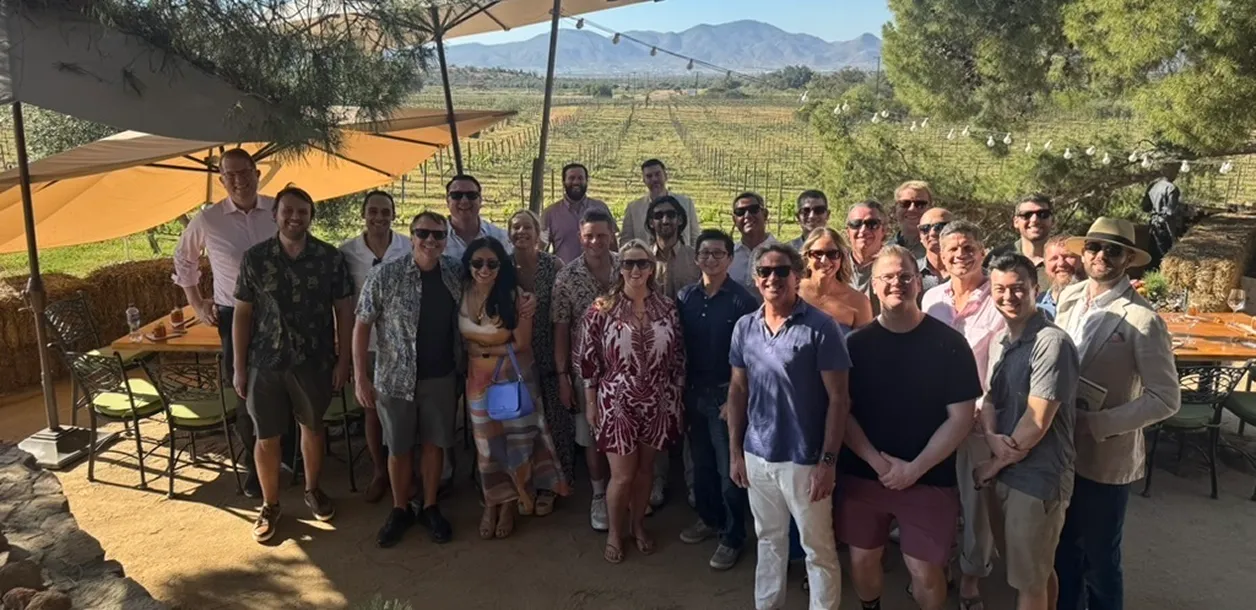 Group of people posing outdoors in front of vineyard under sunny sky with mountains in the background.