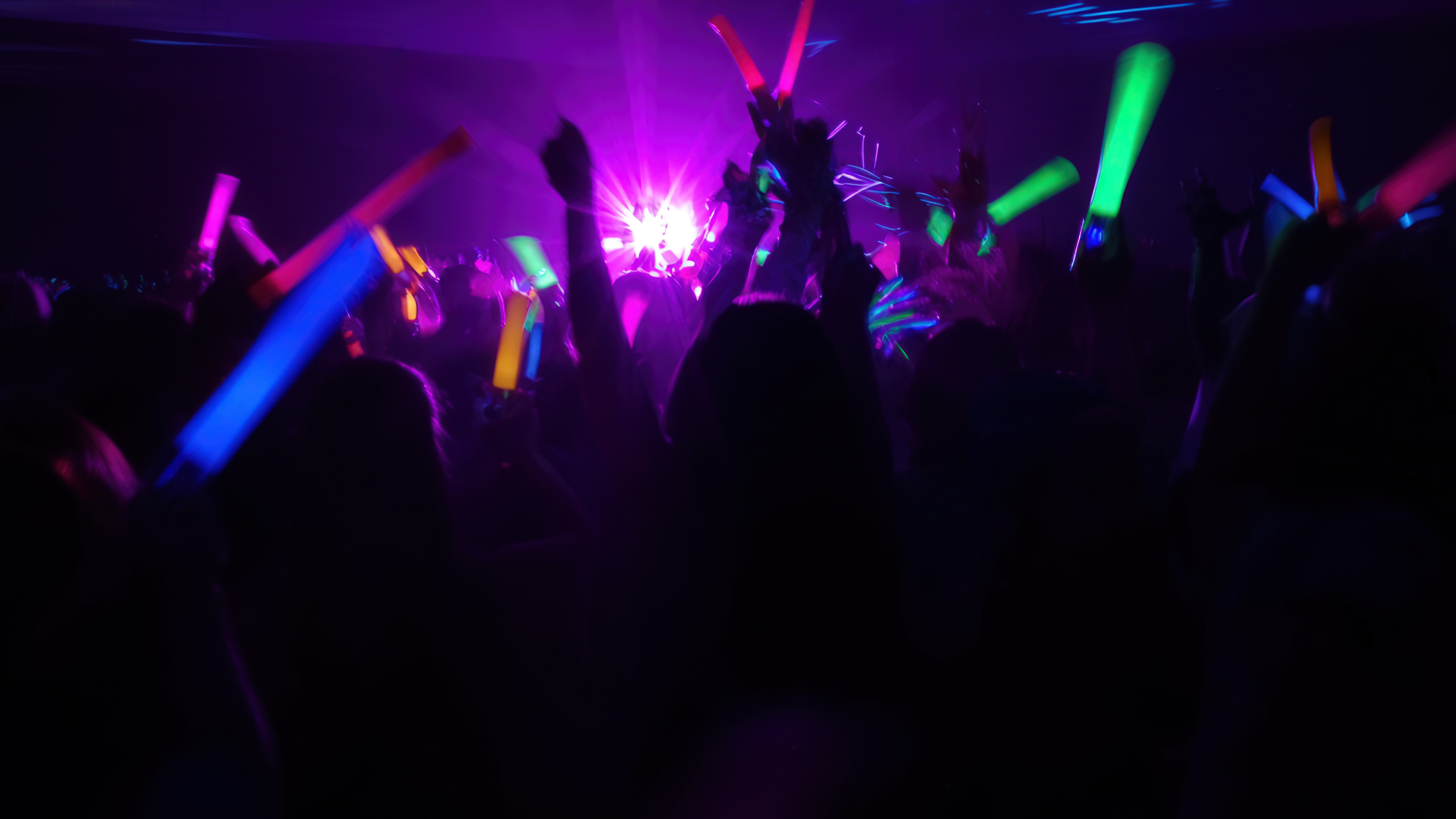 Crowd waving colorful glowing sticks under purple and blue concert lights at a lively event.