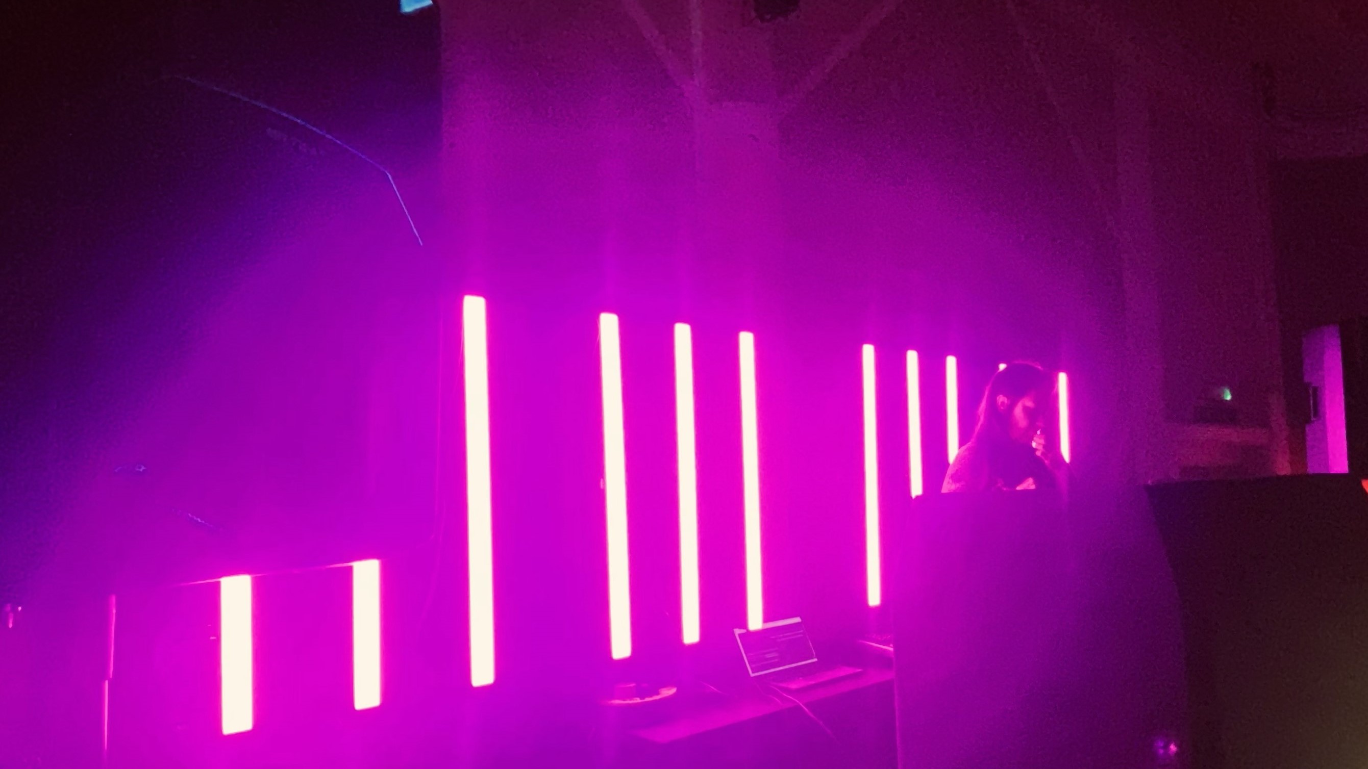 Dimly lit scene with bright vertical pink neon lights and a person working near a laptop in a dark room.