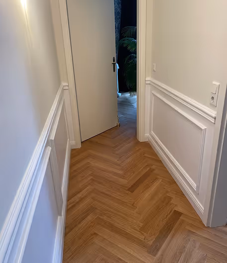 Heller Flur mit weißer Wandverkleidung und Holzboden, offene Tür führt zu einem Raum mit Pflanzen.