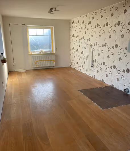 Empty room with wooden floor, floral wallpaper, window covered with plastic, and patched floor area.