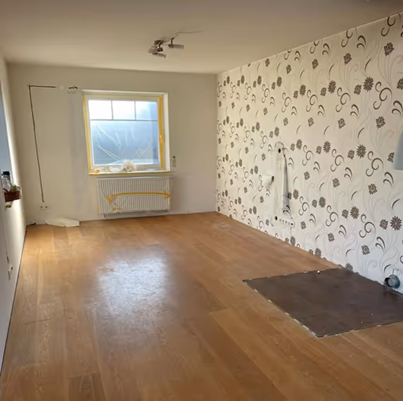 Empty room with wooden floor, floral wallpaper on one wall, a window with taped plastic, and a radiator.