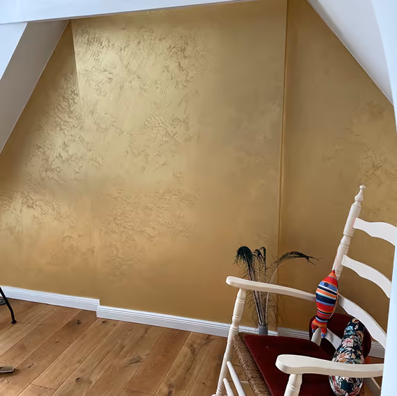 Weißer Holzstuhl mit roten Kissen und Dekoration vor einer goldfarbenen Wand auf Holzboden.