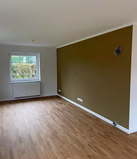 Leerer Raum mit Holzboden, weißer und olivgrüner Wand, Fenster und Heizkörper.