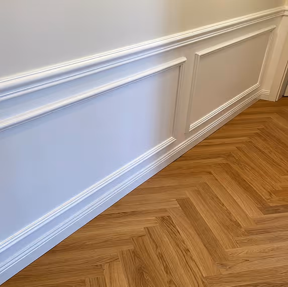 Weiß gestrichene Wand mit dekorativer Leiste über einem Holzboden in Fischgrätmuster.
