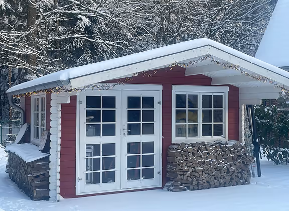 Rotes Gartenhaus mit weißen Türen und Fensterläden, Schnee auf Dach und umgebendem Boden.