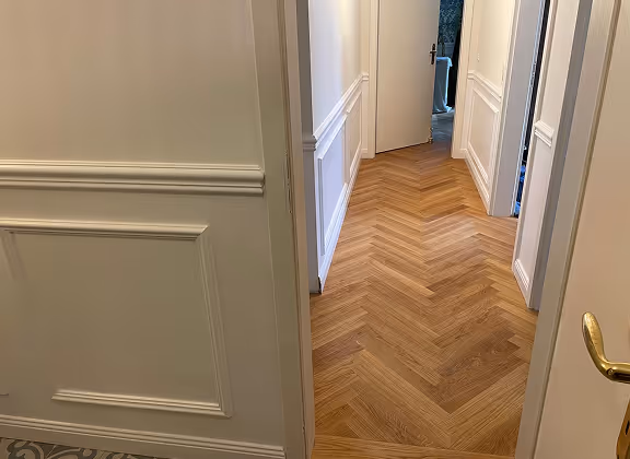 Flur mit weißen Wänden, Holzfußboden im Fischgrätmuster und einer offenen Tür am Ende.