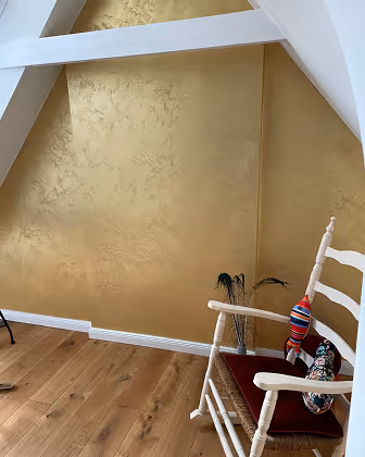 Weißer Holzstuhl mit bunten Kissen vor goldener Wand und Holzboden in einem Raum mit Dachschräge.
