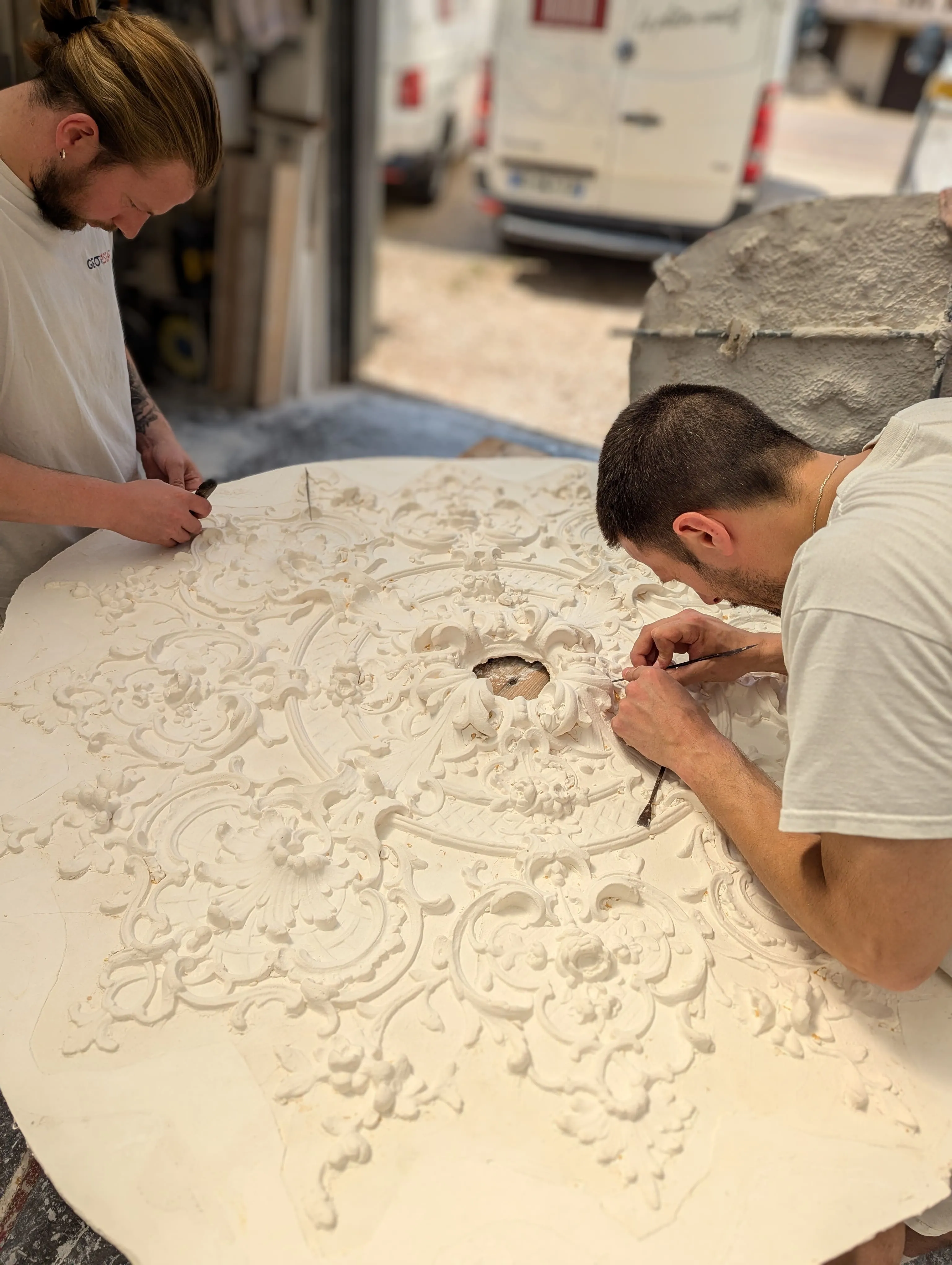 Deux artisans retouchent à la main une grande rosace en plâtre à l'atelier