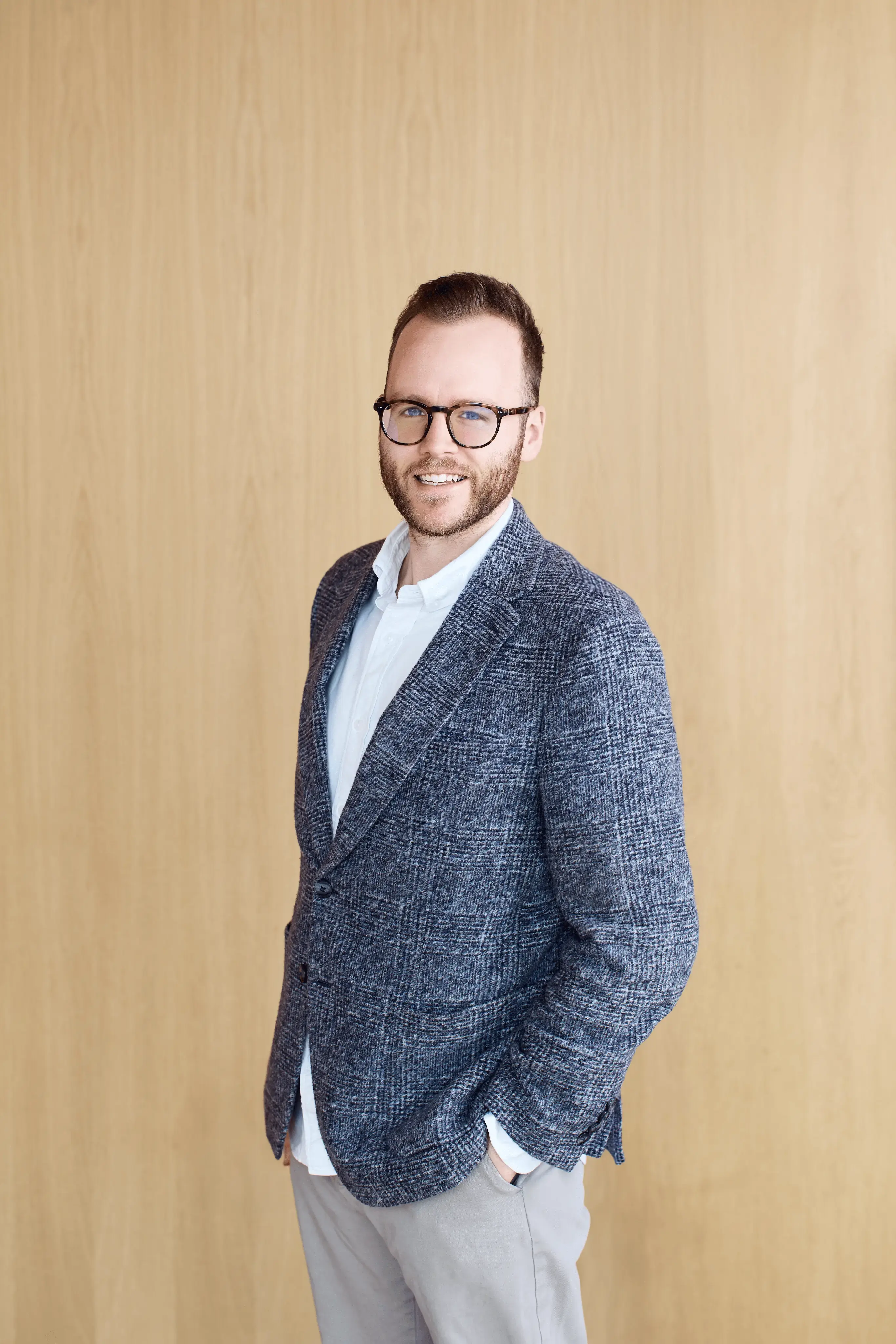 Smiling man with glasses in a gray blazer and white shirt standing against a light wooden wall.