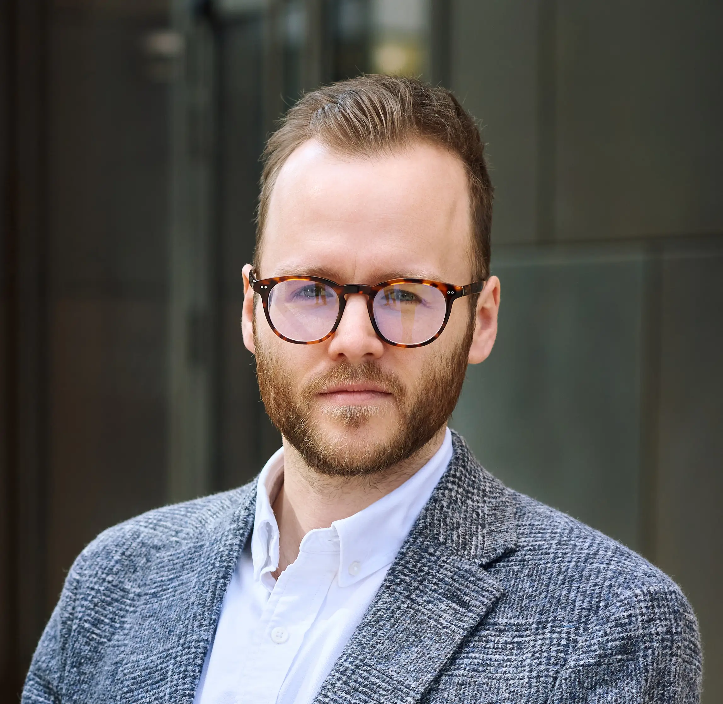 Man with short hair and beard wearing glasses, a white shirt, and a gray plaid blazer.