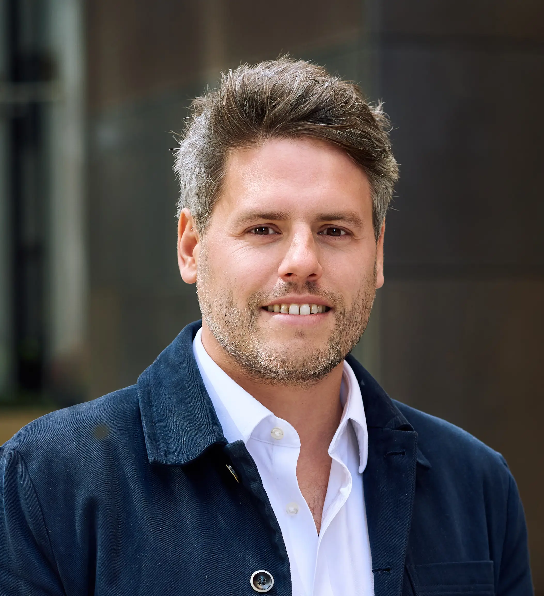 Smiling man with short hair and beard wearing a white shirt and dark jacket in an outdoor setting.