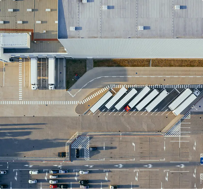 Aerial view of trucks parked at warehouse loading docks and cars lined up in a parking area.