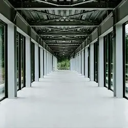 Empty modern corridor with metal beams, glass walls, and a view of trees at the end.