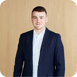 Young man with short hair wearing a navy blazer and white shirt standing in front of a wood wall.