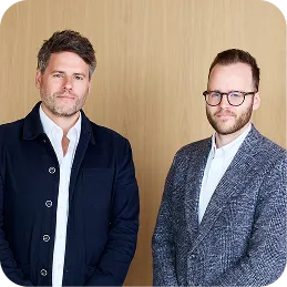 Two men standing in front of a wooden wall, one in a navy jacket and the other in a gray blazer with glasses.