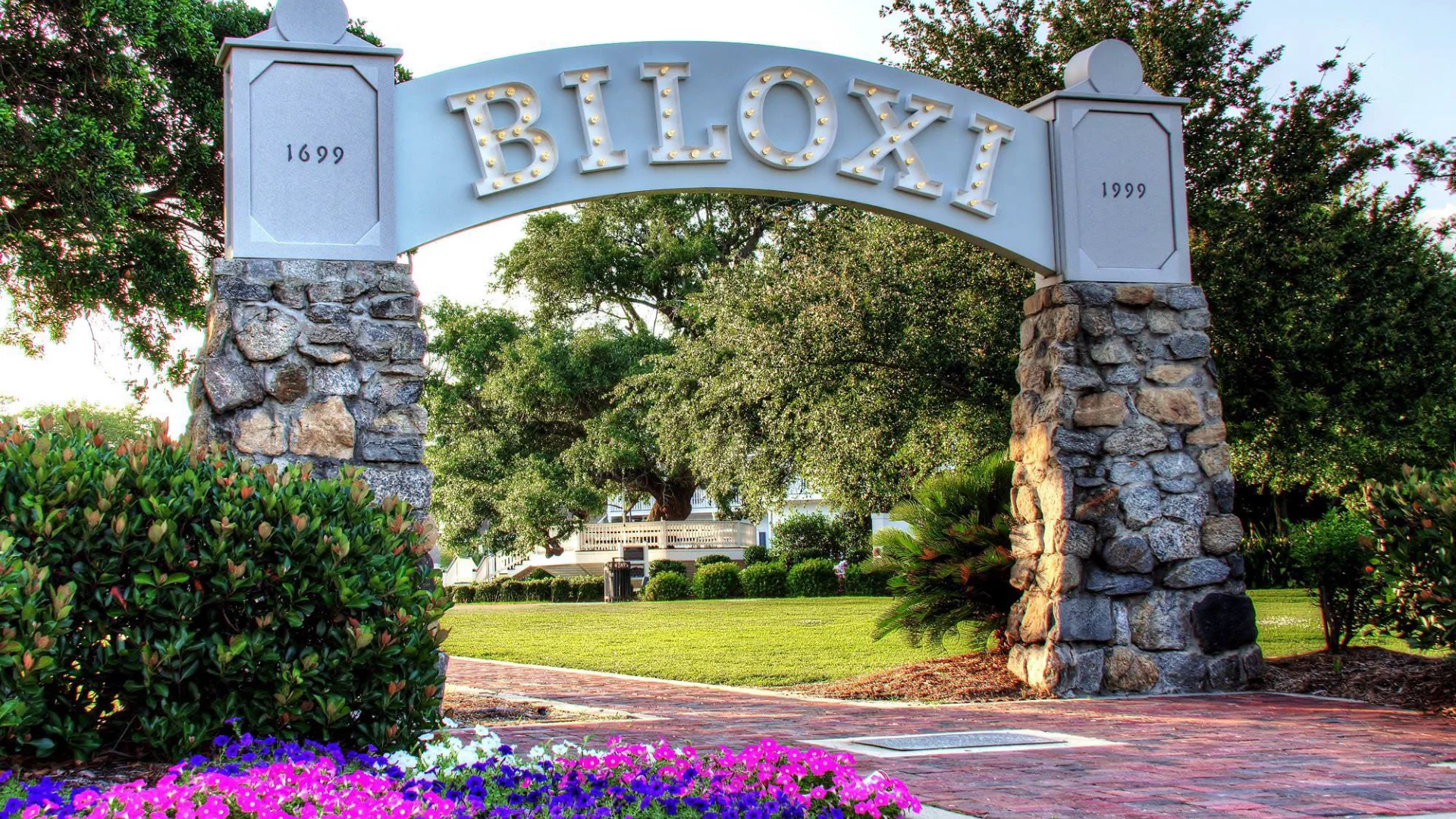 Stone archway with lit letters spelling BILOXI over a brick path with flowers and greenery around.