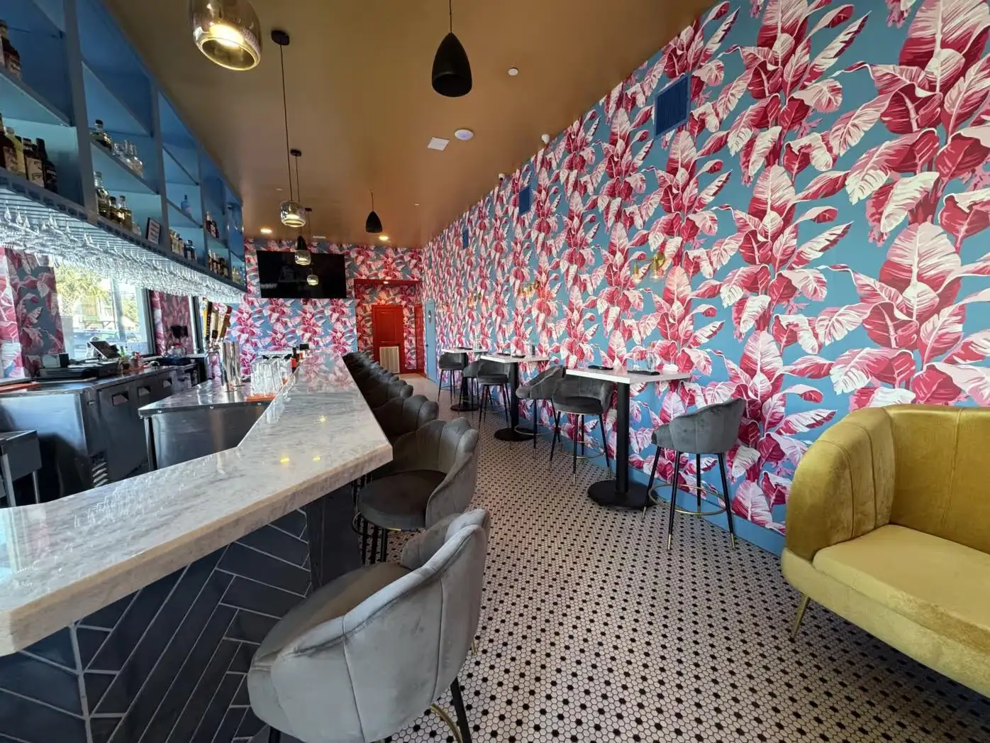 Modern bar interior with floral wallpaper, marble countertop, grey bar stools, and patterned tile floor.