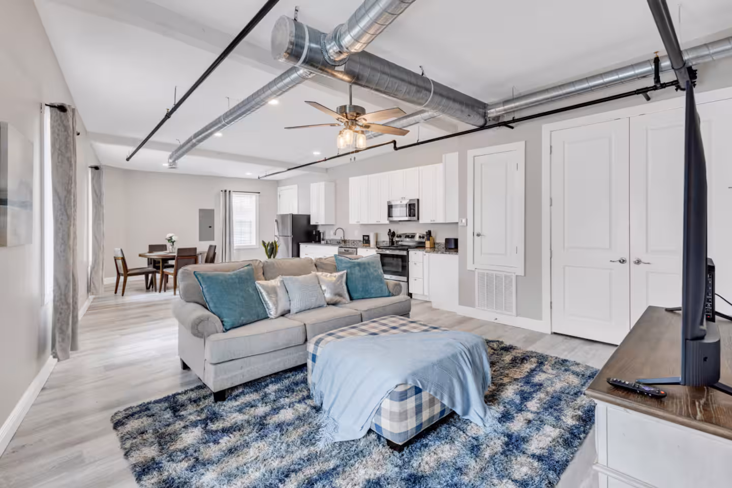 Modern open living room with gray sofa, blue pillows, checked ottoman, and kitchen in the background.