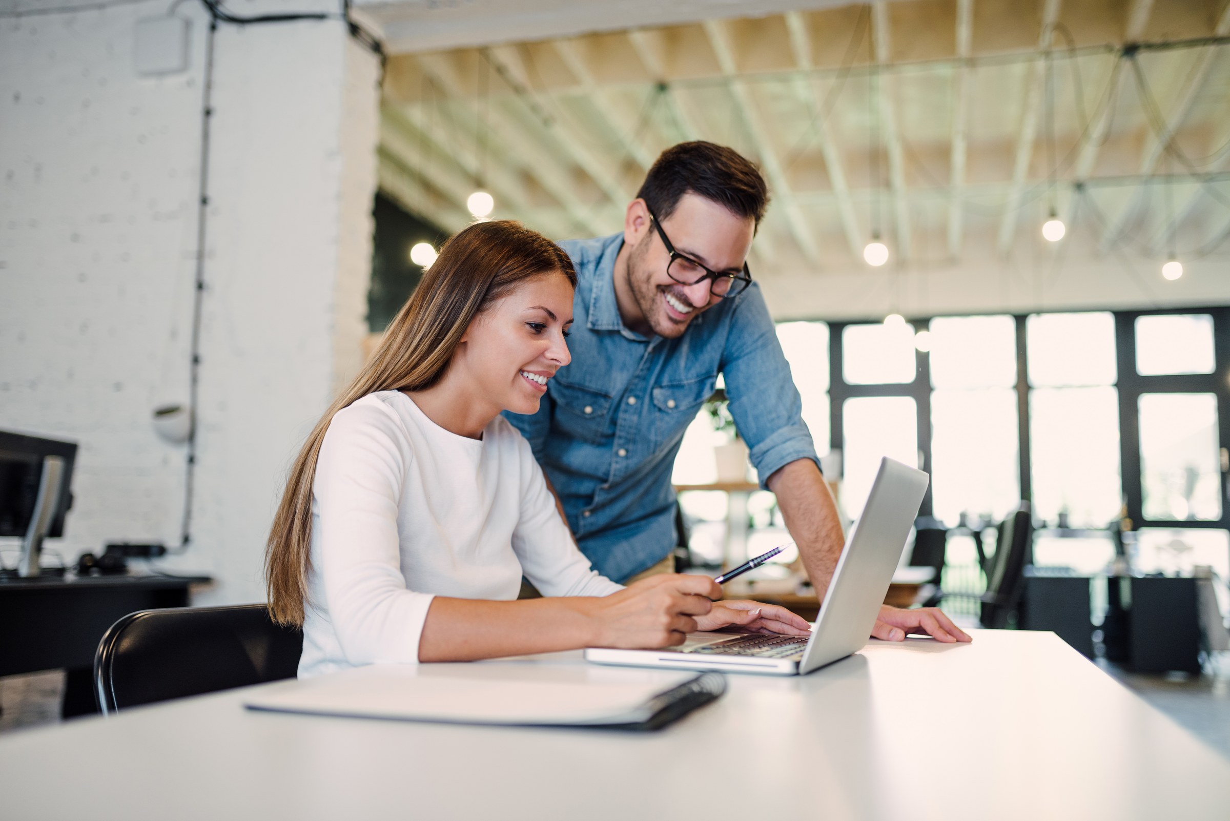 Two coworkers smile as they work together on a laptop, with one taking notes beside it.