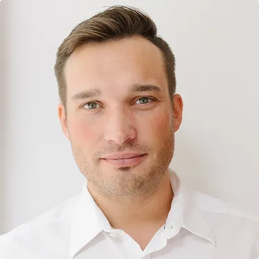 Mann mit kurzen braunen Haaren, grün-braunen Augen und weißem Hemd vor hellem Hintergrund.