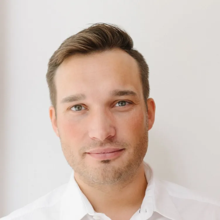 Mann mit kurzen braunen Haaren, grün-braunen Augen und weißem Hemd vor hellem Hintergrund.