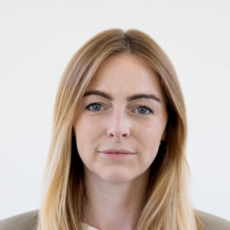 Porträt einer Frau mit langen blonden Haaren und blauem Blick, die in grauem Blazer und weißem Hemd schaut.