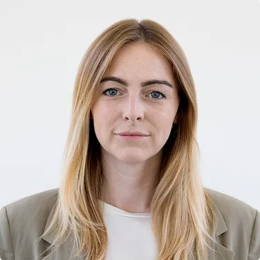 Porträt einer Frau mit langen blonden Haaren und blauem Blick, die in grauem Blazer und weißem Hemd schaut.