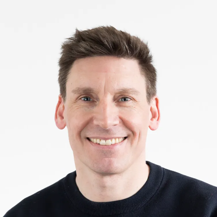 Portrait eines lächelnden Mannes mit dunklem Pullover vor weißem Hintergrund.