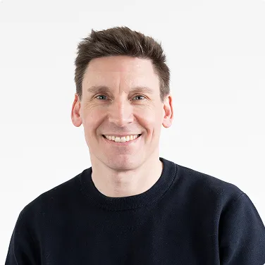 Portrait eines lächelnden Mannes mit dunklem Pullover vor weißem Hintergrund.