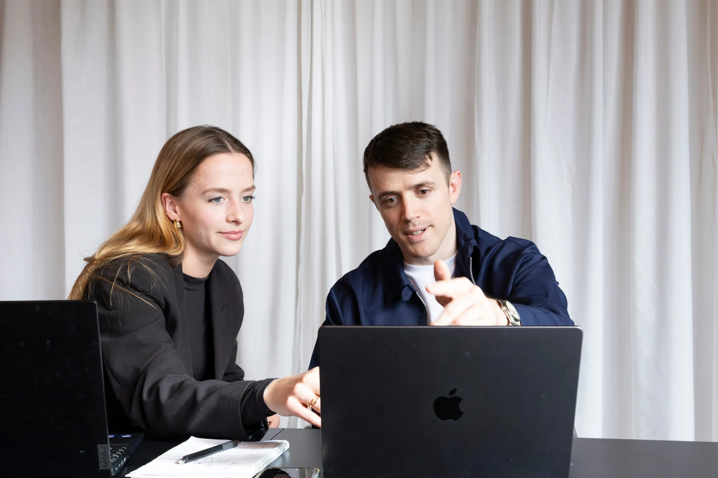 A man and a woman are working together on a laptop in front of a white curtain.