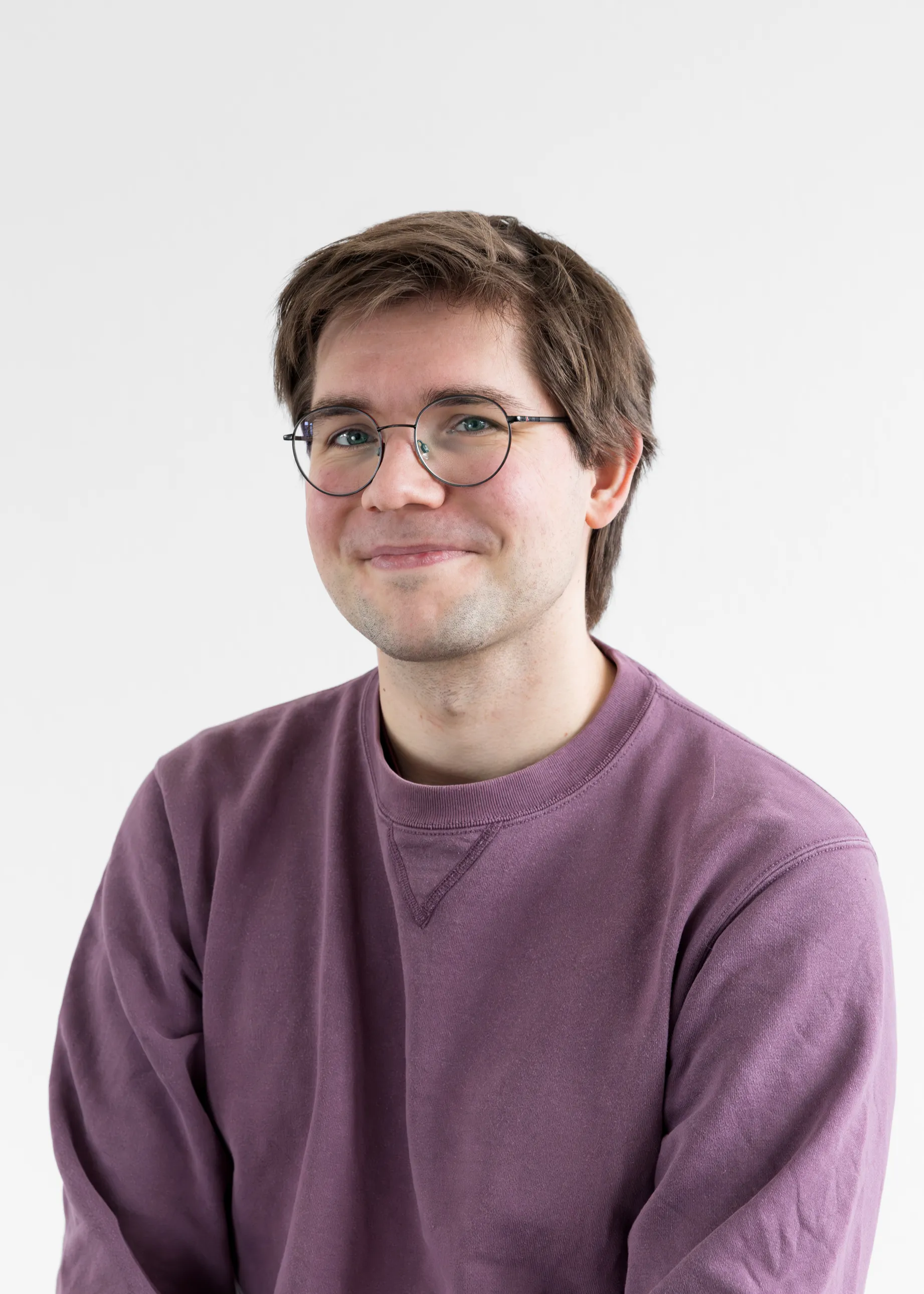 Junger Mann mit braunen Haaren, Brille und einem violetten Pullover vor weißem Hintergrund.