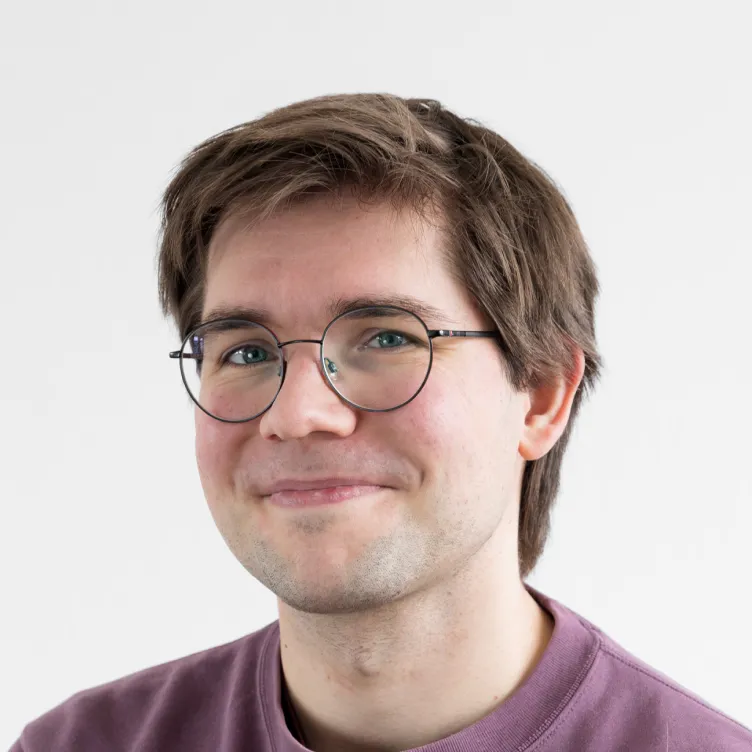 Junger Mann mit braunen Haaren, Brille und einem violetten Pullover vor weißem Hintergrund.