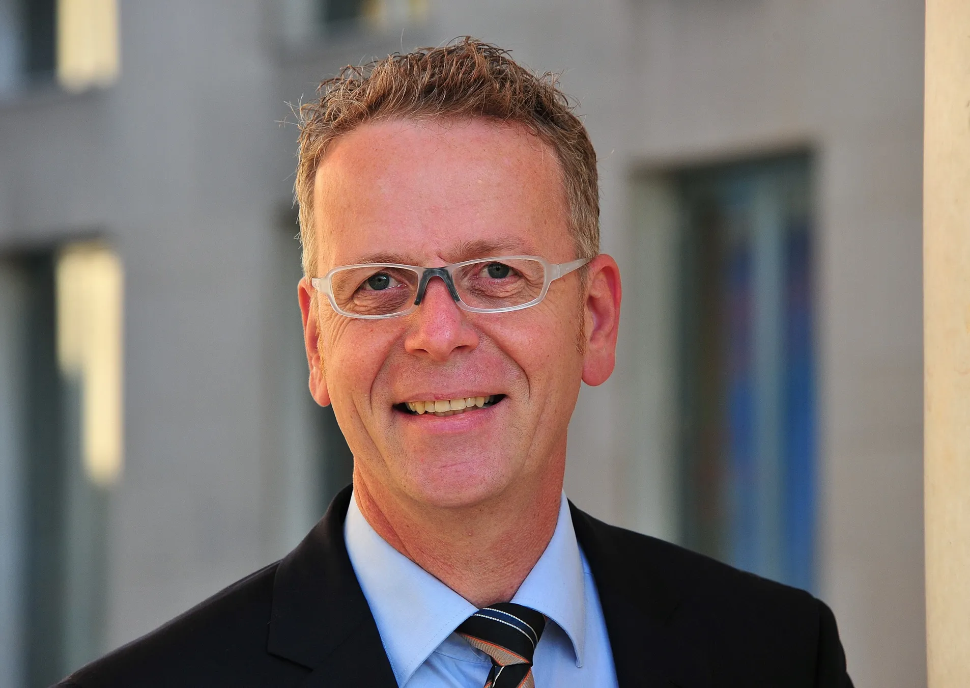 Porträt eines lächelnden Mannes mit Brille in Anzug und Krawatte vor unscharfem Hintergrund.