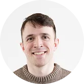 Portrait of a smiling young man with short brown hair and a brown sweater.