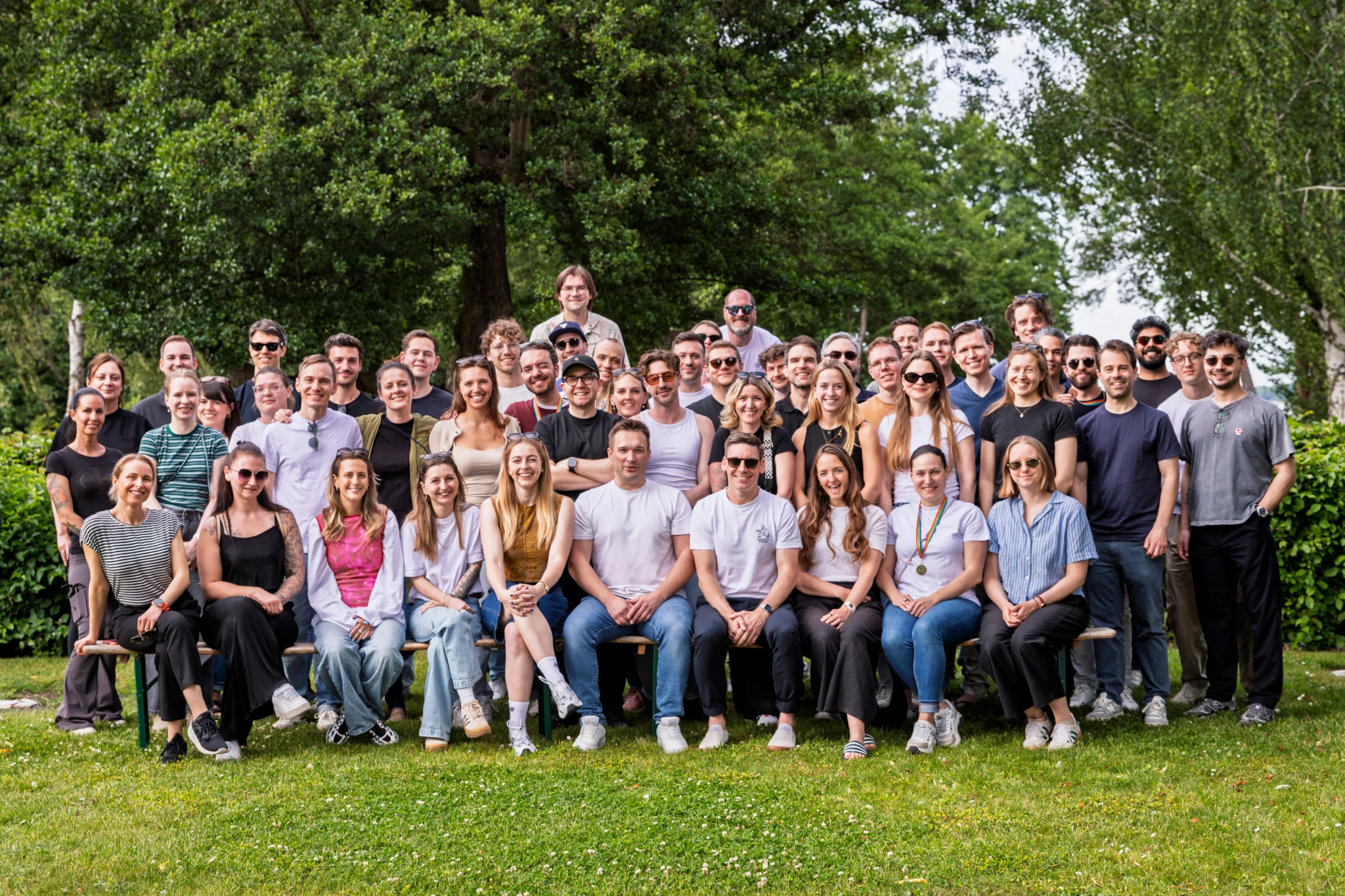 Großes Team von etwa 50 Personen sitzt und steht auf Wiese vor grünen Bäumen, lächelnd.