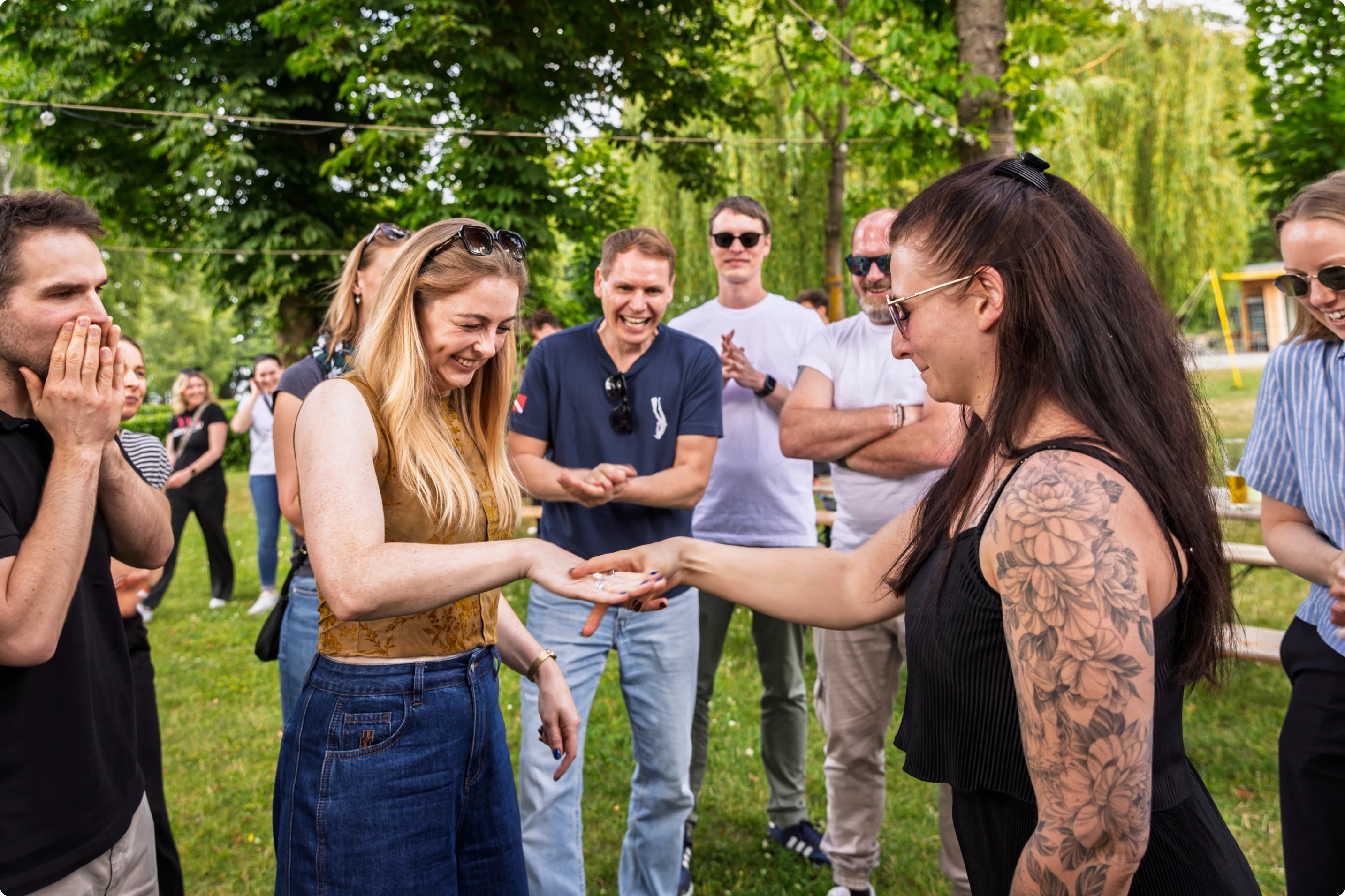 Frauen lachen und zeigen Hände bei einer Outdoor-Aktivität, umgeben von Menschen im Garten.