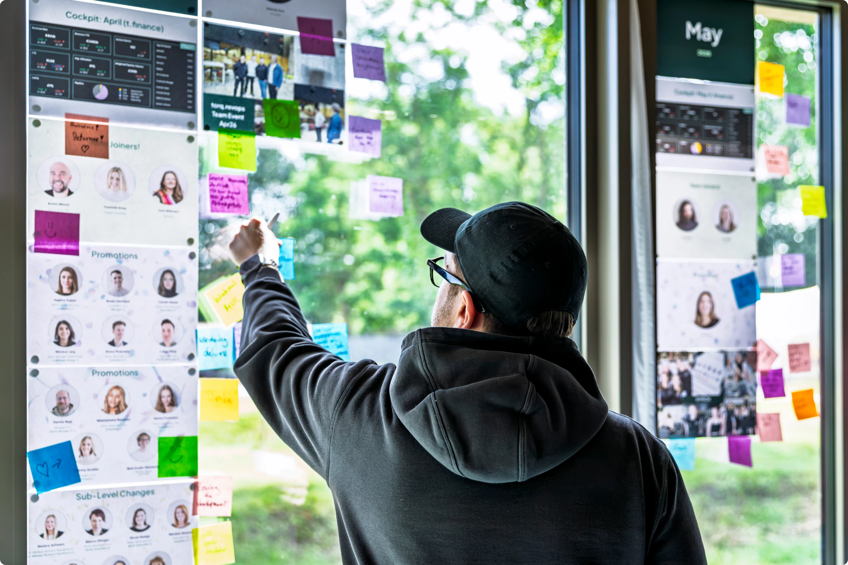 Person wearing a cap and hoodie arranges colorful sticky notes on a glass wall with papers posted.