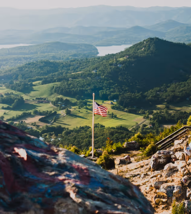 American flag on a pole overlooking a scenic valley with green hills, trees, and a lake in the distance under a hazy sky.