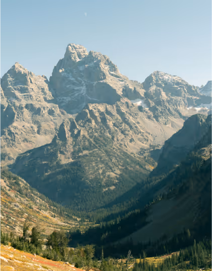 Large rugged mountain range with rocky peaks and forested valley under clear blue sky.