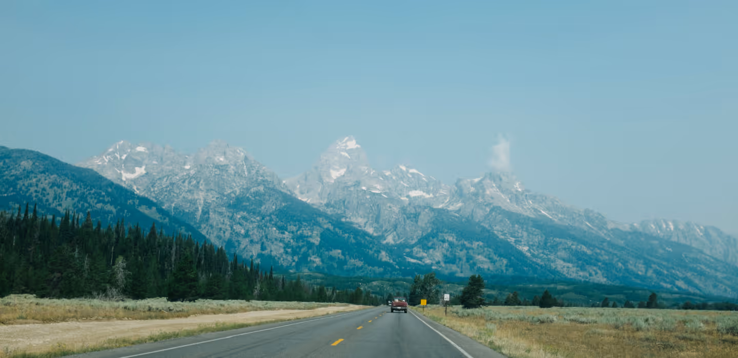 Open road leading towards tall, snow-capped mountains with a car driving ahead under clear blue sky.