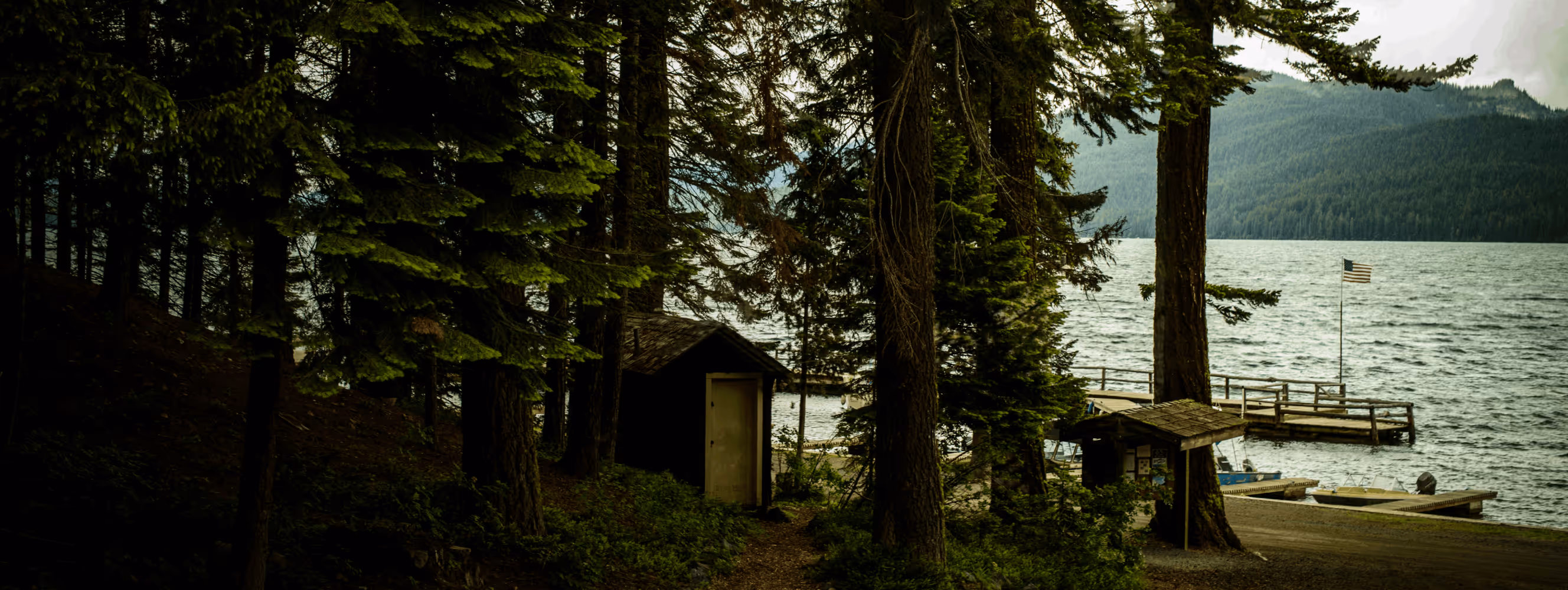 A lakeside scene with tall evergreen trees, a small cabin with a closed door, wooden docks extending into the water, and an American flag on a flagpole near the shore.