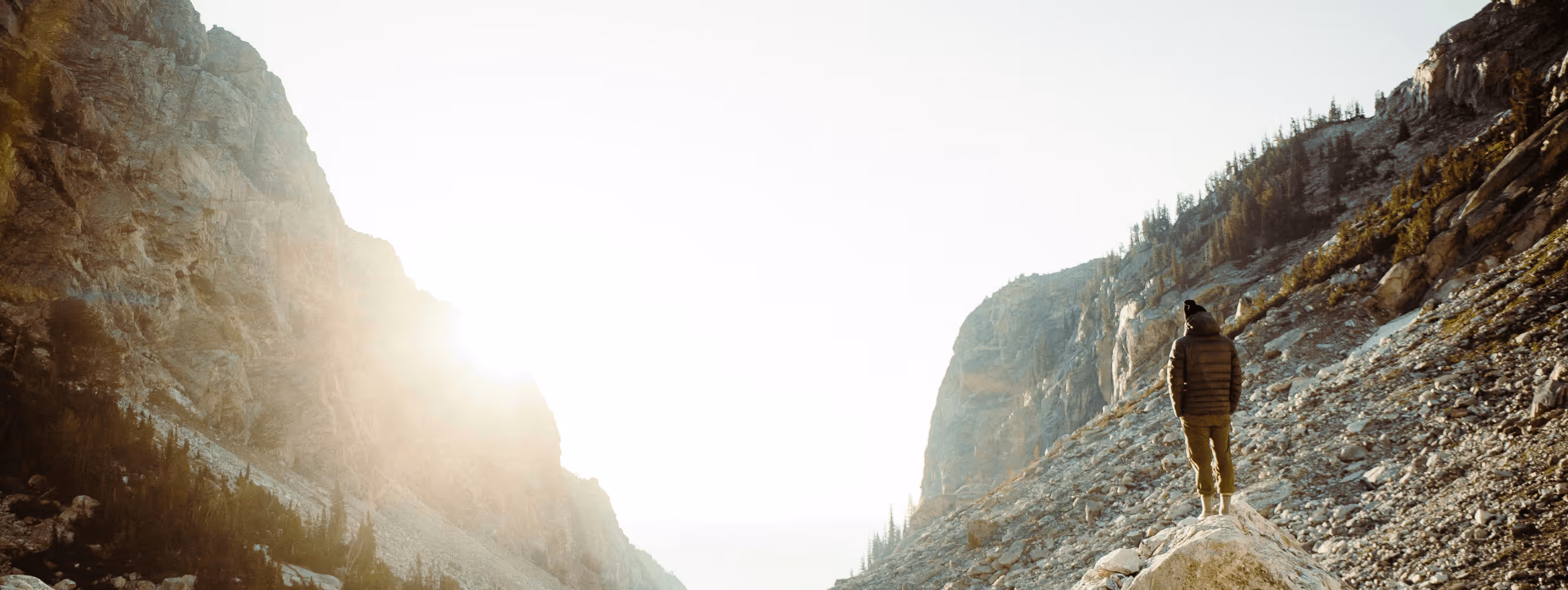 Person in outdoor gear standing on a rock looking at tall rocky mountain cliffs with sunlight shining through.